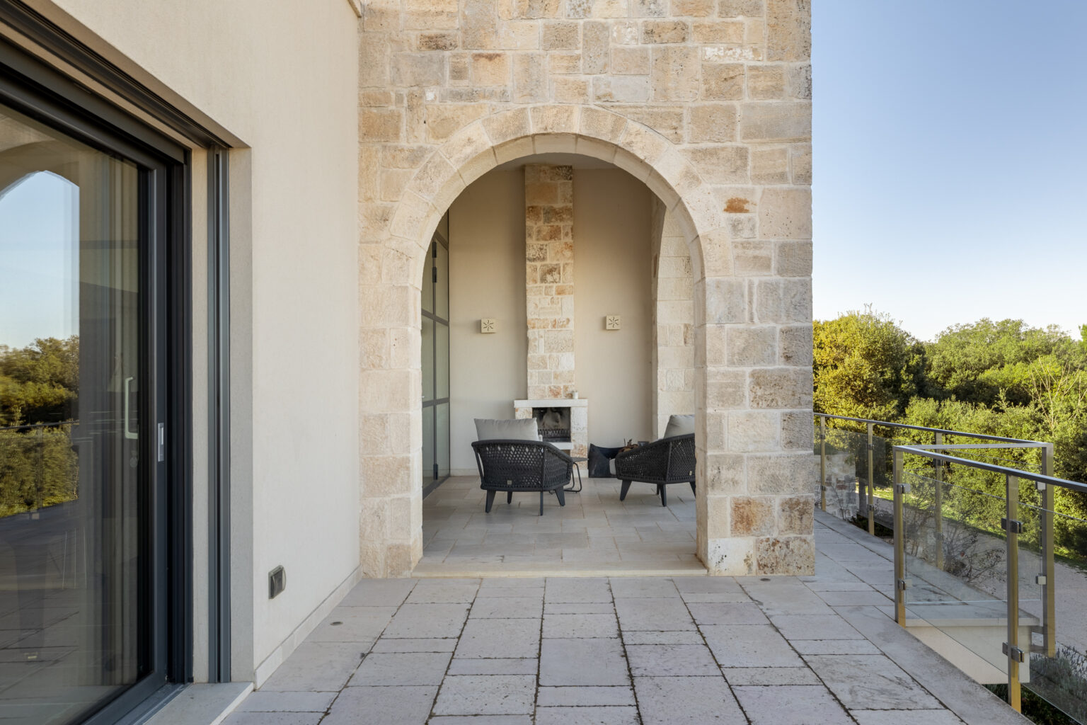 Modern outdoor terrace with a stone archway, featuring two black wicker chairs and a small table, surrounded by lush greenery. | Only on SopranoVillas