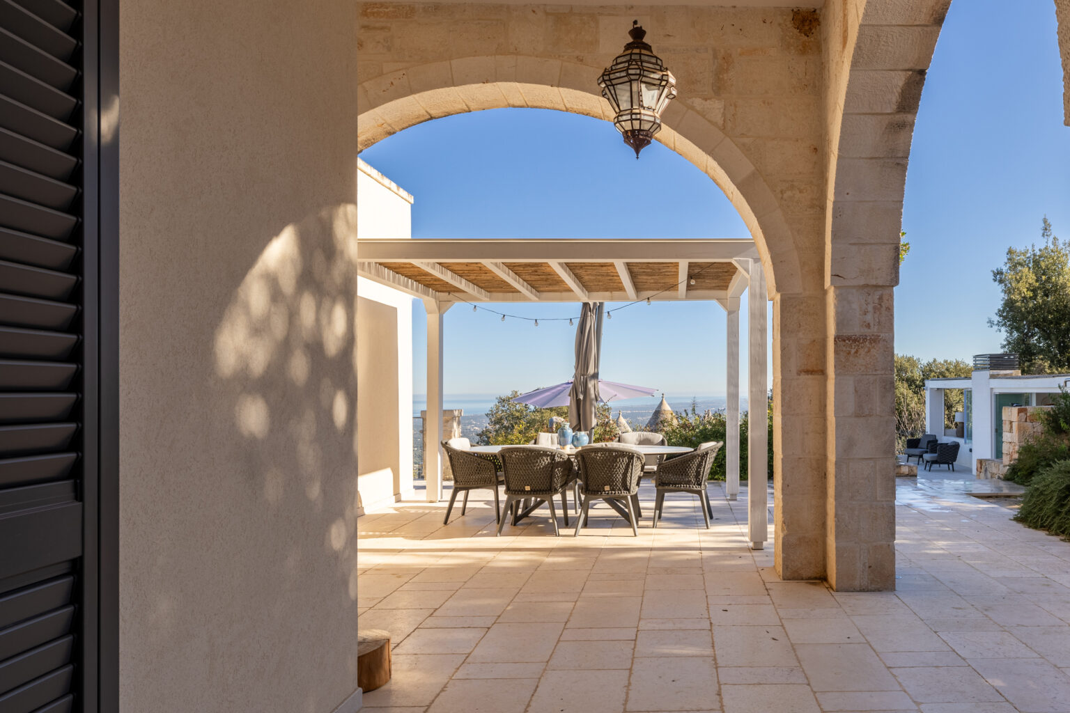 Outdoor dining area under a pergola with a stunning view, featuring a round table and chairs, framed by an elegant stone archway and a hanging lantern. Ideal for entertaining and enjoying nature. | Only on SopranoVillas