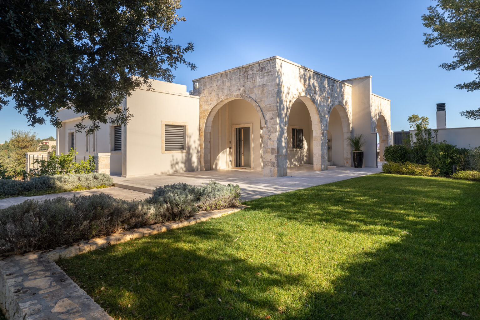 Modern stone villa with arched entryways, surrounded by lush green lawn and trees, under a clear blue sky. | Only on SopranoVillas