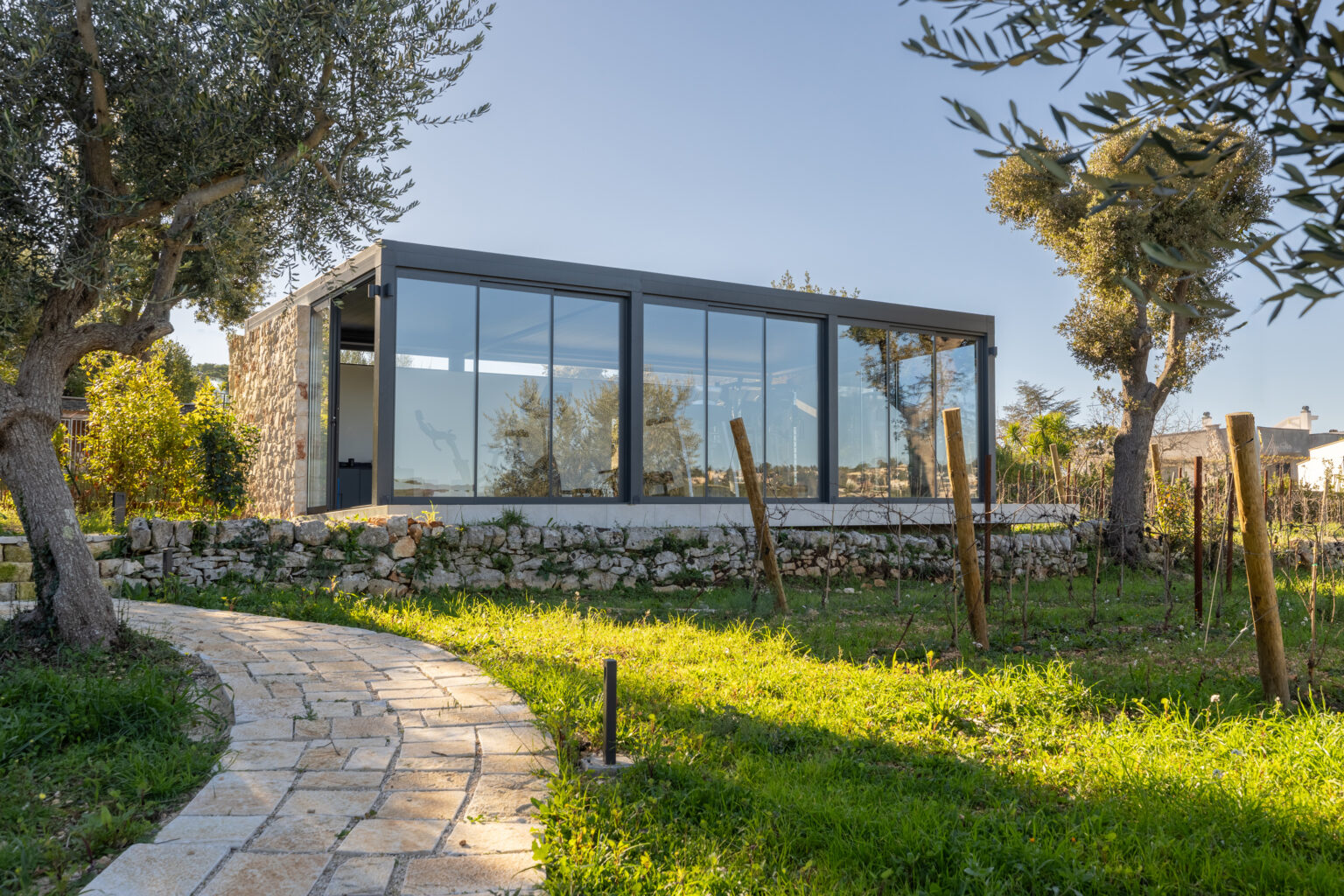 Modern glass house surrounded by greenery and olive trees, featuring a stone pathway leading to the entrance. | Only on SopranoVillas