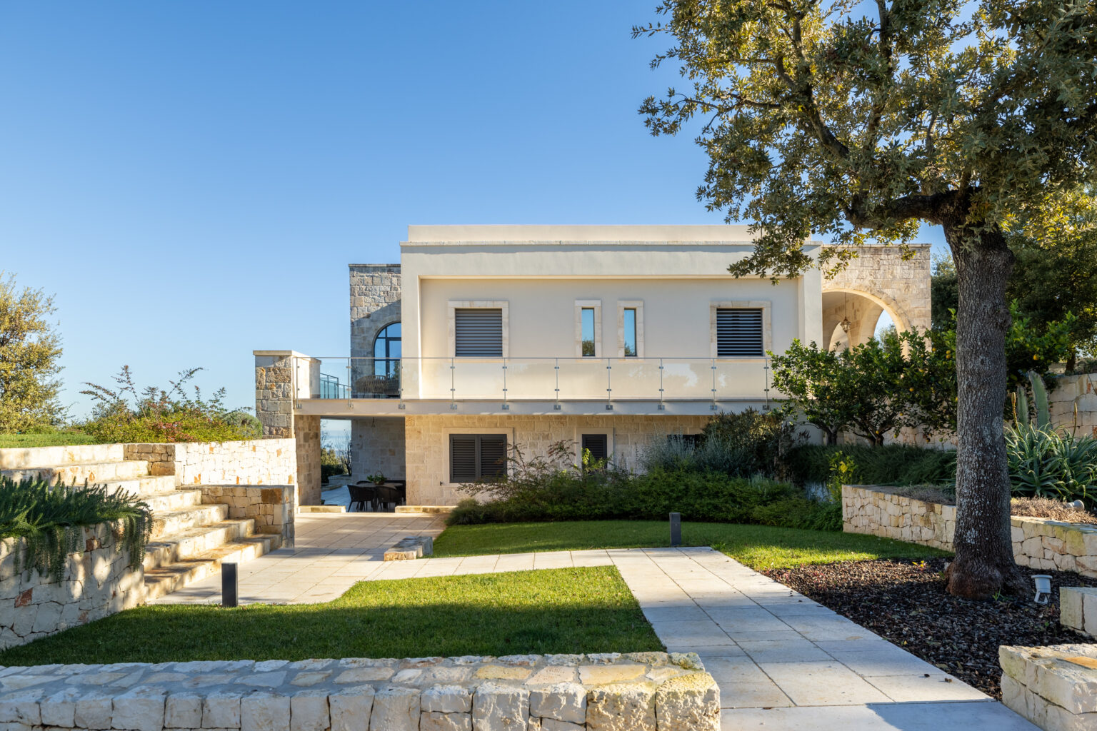 Modern villa with a landscaped garden, stone steps, and a clear blue sky in the background. The architecture features a mix of stone and smooth walls, surrounded by greenery. | Only on SopranoVillas
