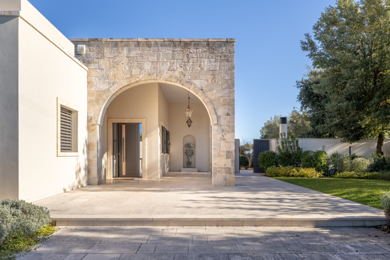 Modern stone house entrance with an arched doorway, surrounded by a landscaped garden and clear blue sky. Ideal for showcasing contemporary architecture and outdoor living spaces. | Only on SopranoVillas