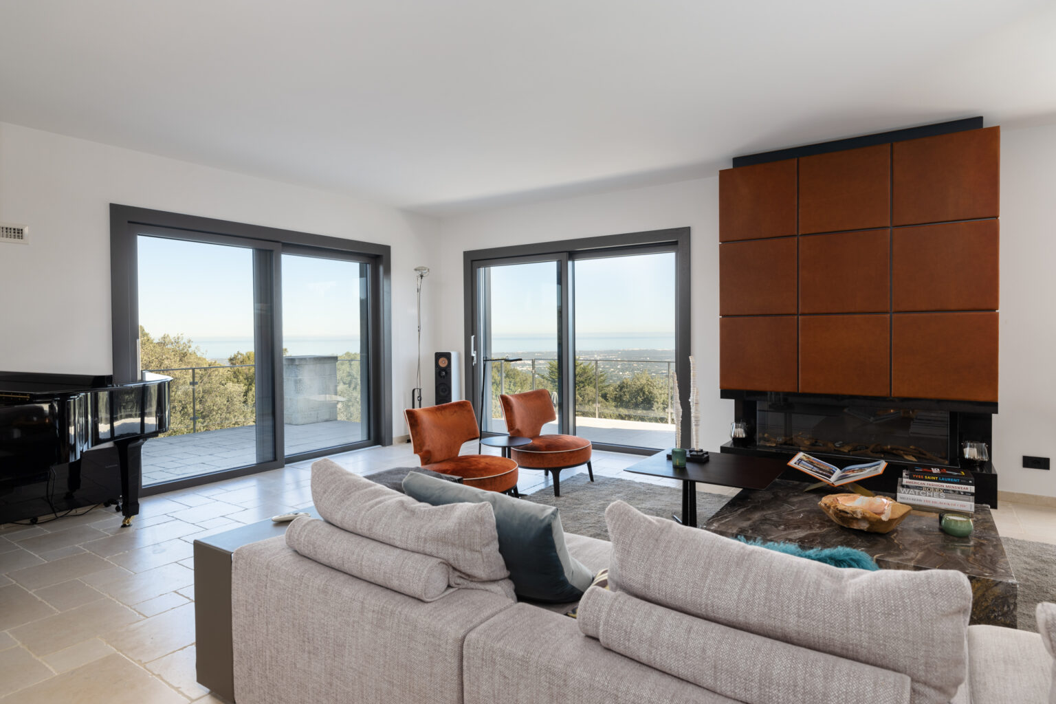Modern living room featuring large windows with scenic views, a comfortable gray sofa, two orange armchairs, a sleek black piano, and a stylish wooden accent wall. Natural light enhances the spacious atmosphere. | Only on SopranoVillas