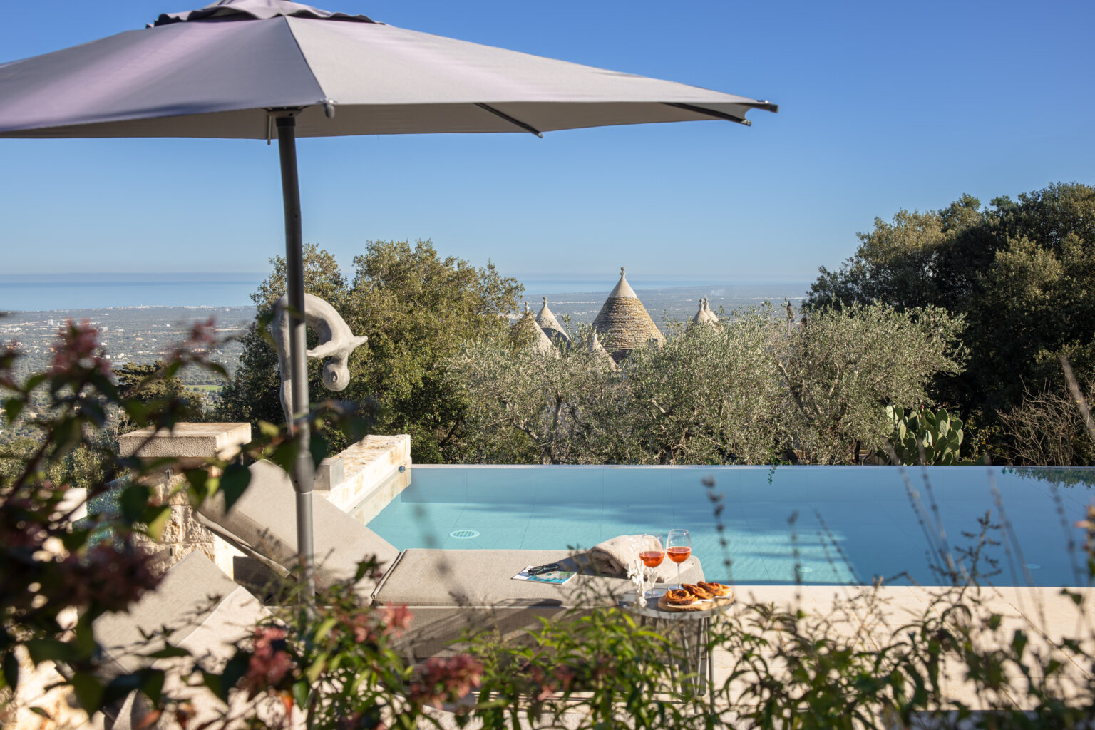 Scenic view of a luxury infinity pool surrounded by lush greenery and traditional trulli houses, with a sun umbrella providing shade, showcasing a tranquil outdoor setting. | Only on SopranoVillas