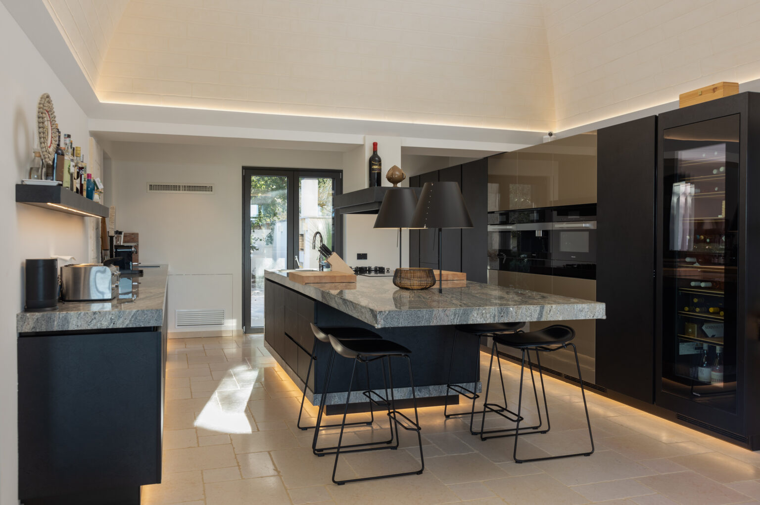 Modern kitchen interior featuring a sleek black design, a large gray marble island with bar stools, stylish pendant lighting, and an open layout that connects to outdoor views. | Only on SopranoVillas
