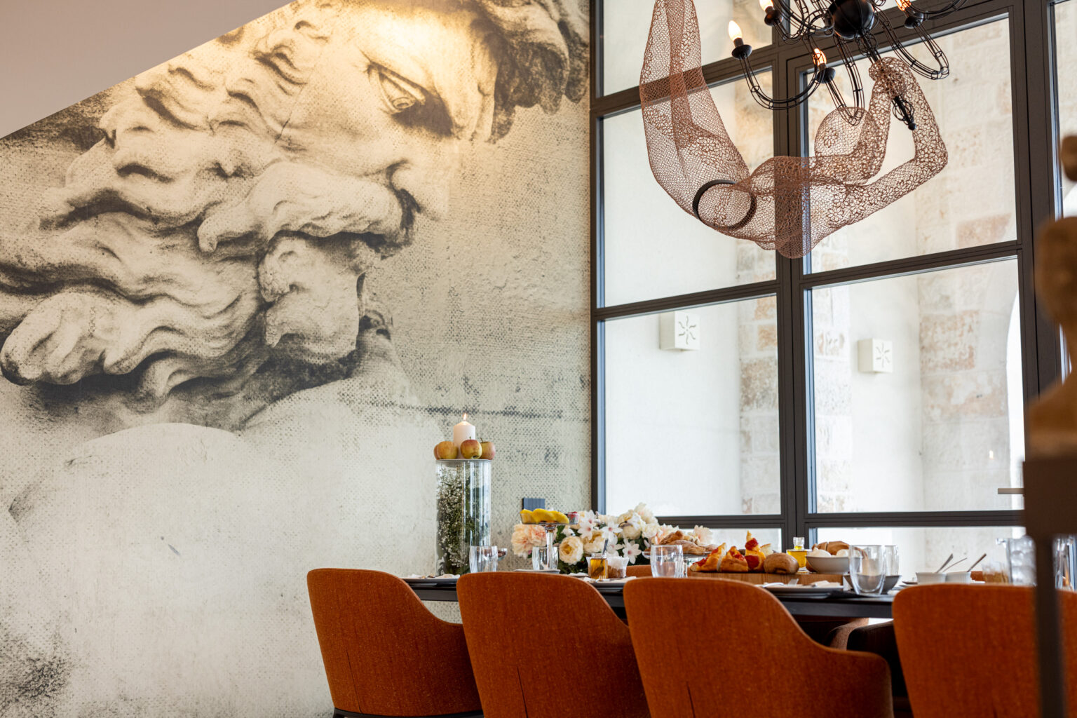 Stylish dining area featuring a modern chandelier, orange upholstered chairs, and a decorative wall with artistic mural elements, creating an inviting atmosphere for meals and gatherings. | Only on SopranoVillas
