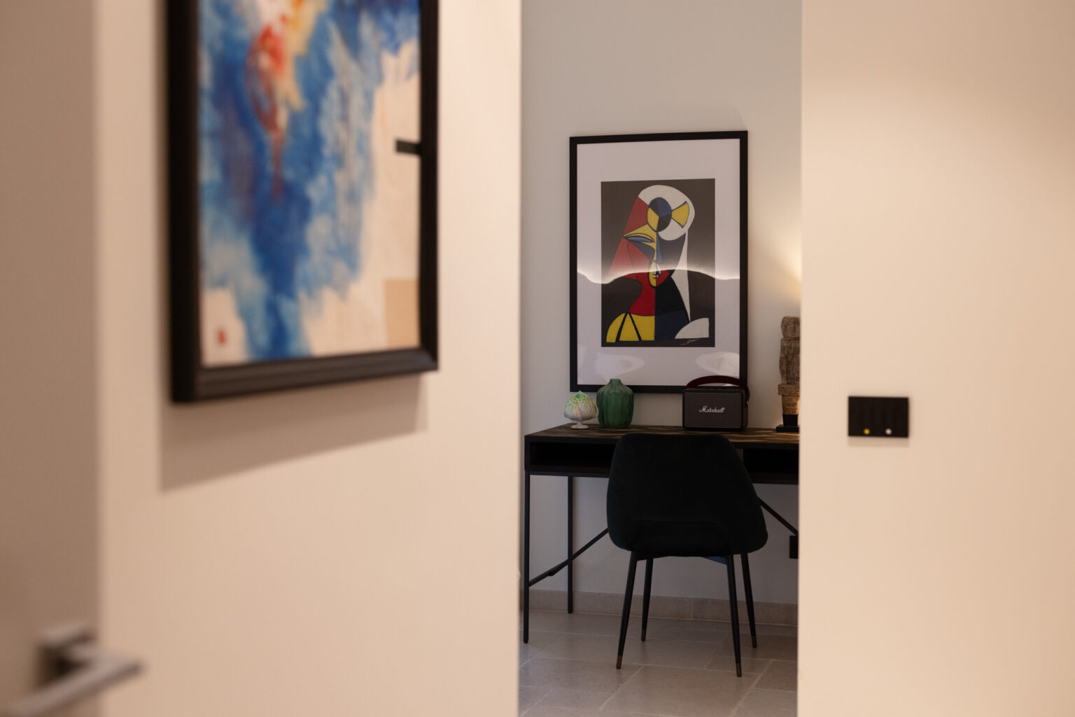 Interior view of a modern workspace featuring a stylish black chair and a sleek desk with decorative items, including a green vase and a Marshall speaker, framed by a doorway. The wall displays colorful artwork, adding a contemporary touch to the room's design. | Only on SopranoVillas