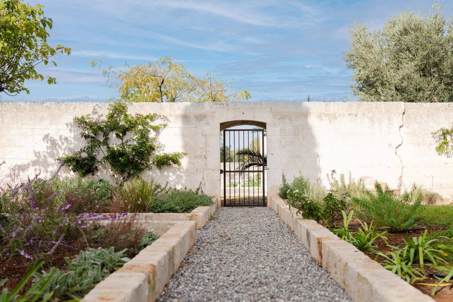 Rustic garden pathway leading to a wrought iron gate, surrounded by lush greenery and flowering plants, with a textured stone wall and blue sky in the background. | Only on SopranoVillas