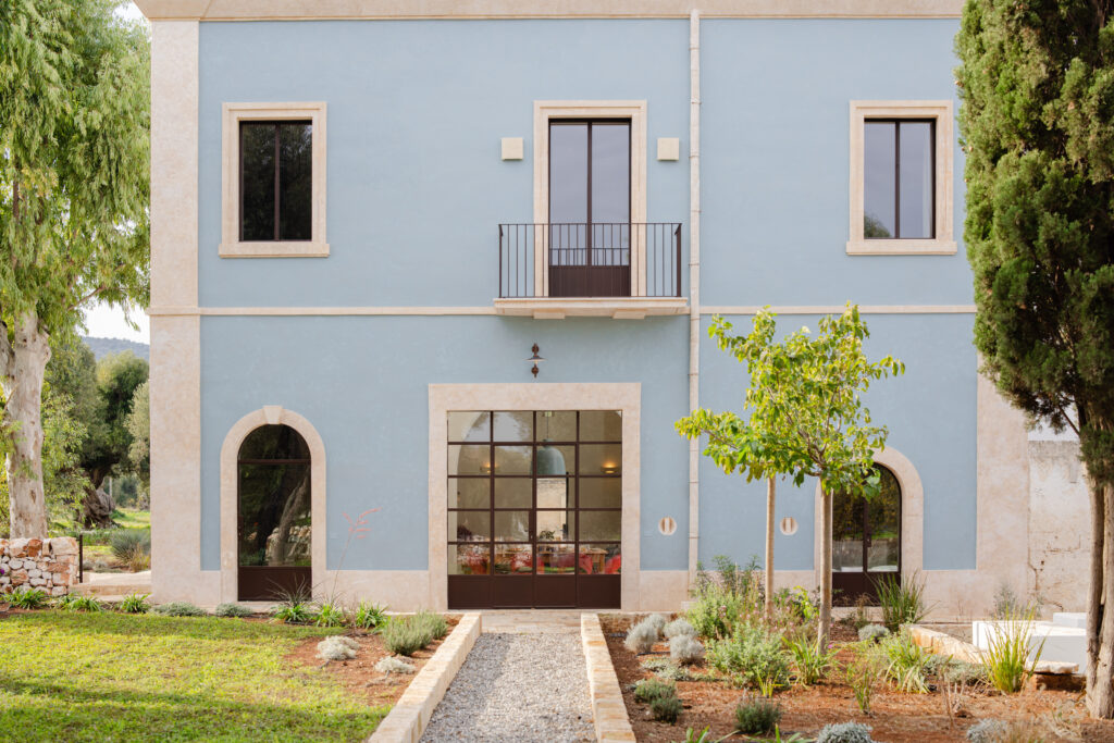Charming blue exterior of a modern villa featuring large windows, a balcony, and landscaped gardens with various plants and gravel pathways, set against a serene natural backdrop. | Only on SopranoVillas
