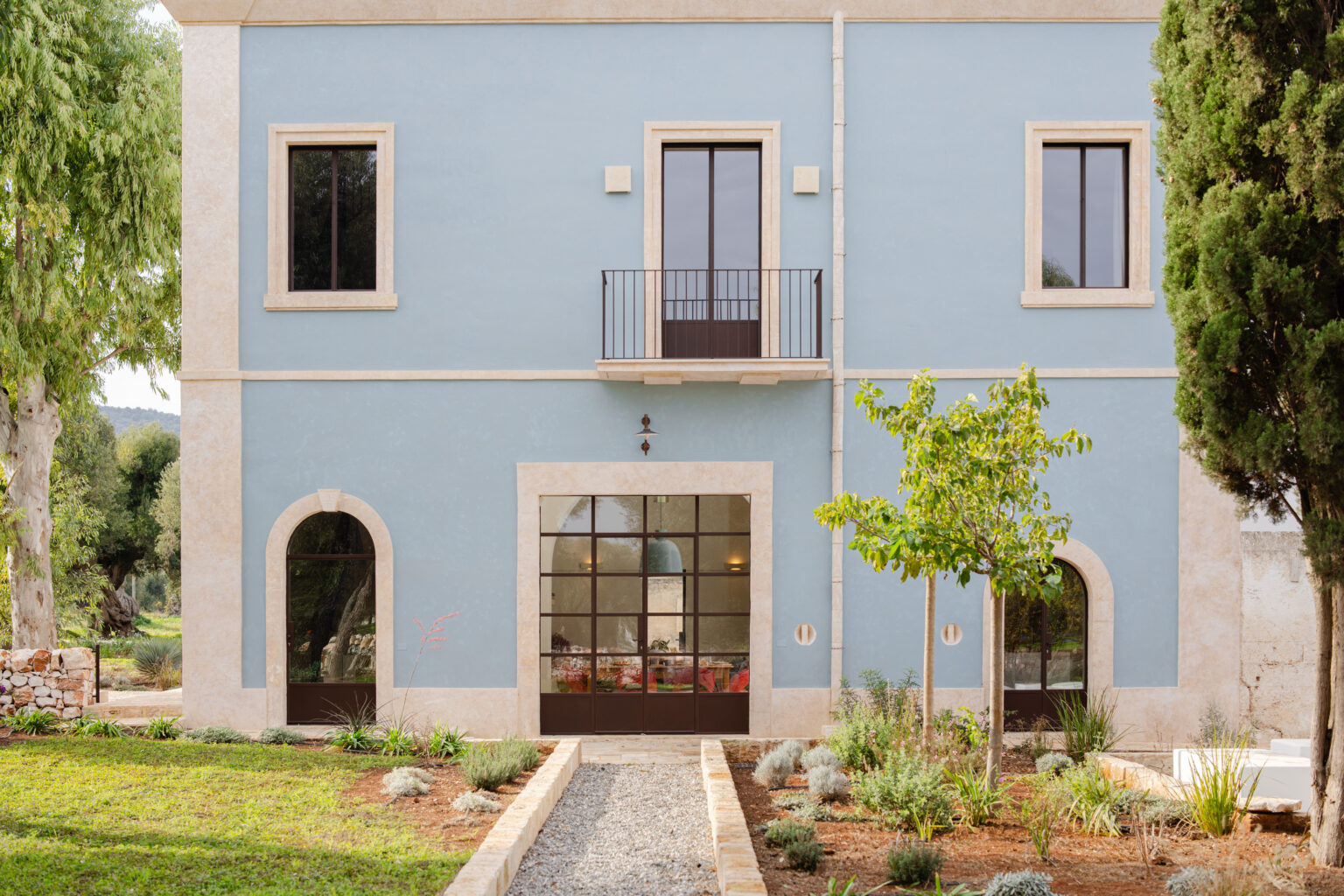 Charming blue exterior of a modern villa featuring large windows, a balcony, and landscaped gardens with various plants and gravel pathways, set against a serene natural backdrop. | Only on SopranoVillas