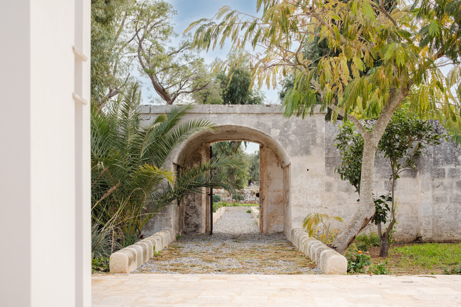 A stone archway leads to a serene garden pathway surrounded by lush greenery, featuring palm trees and a mix of vibrant plants, set against a backdrop of weathered stone walls. | Only on SopranoVillas