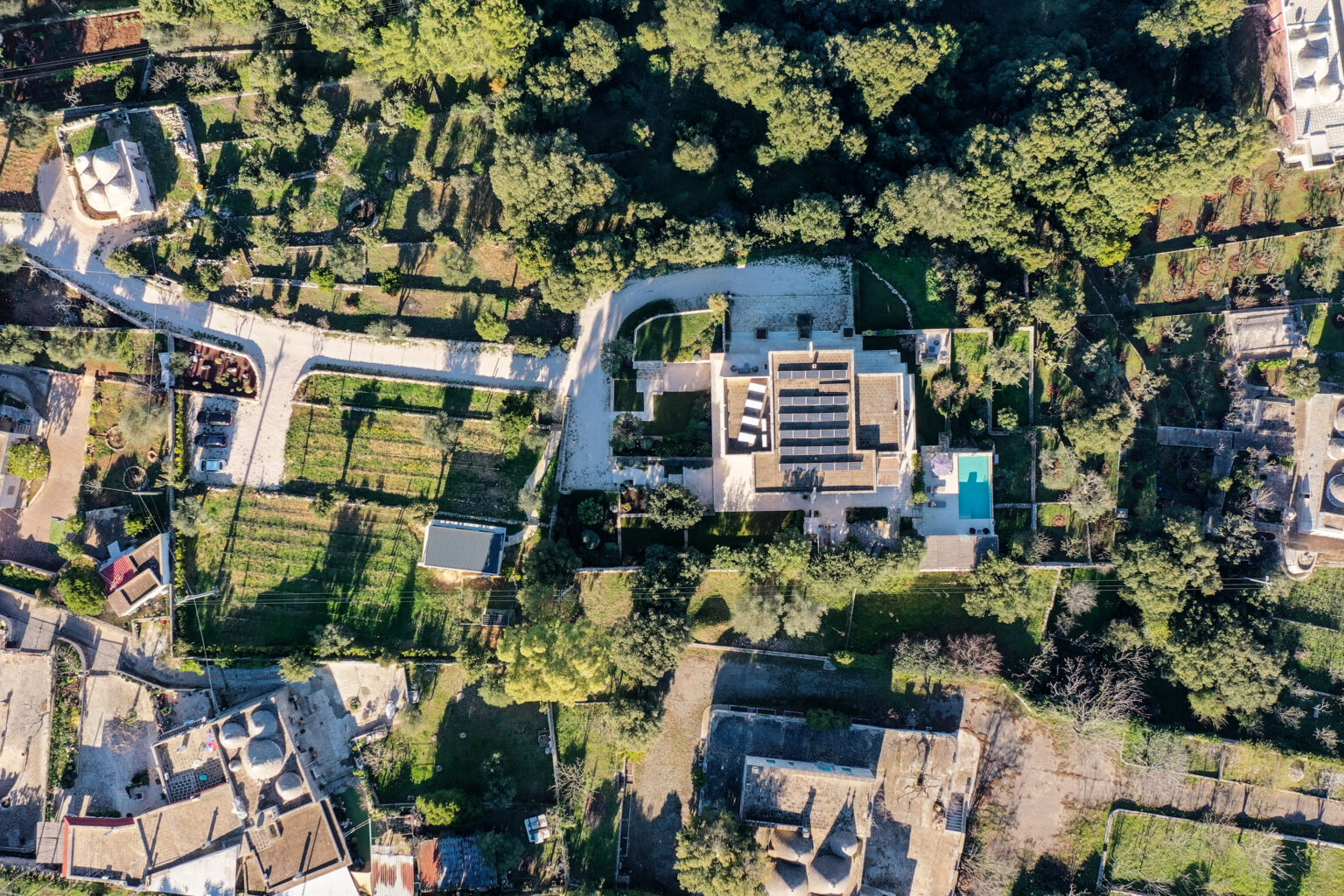 Aerial view of a modern villa surrounded by lush greenery and landscaped gardens, featuring a swimming pool and solar panels on the roof. The image showcases nearby roads and various residential structures in a tranquil, nature-rich environment. | Only on SopranoVillas