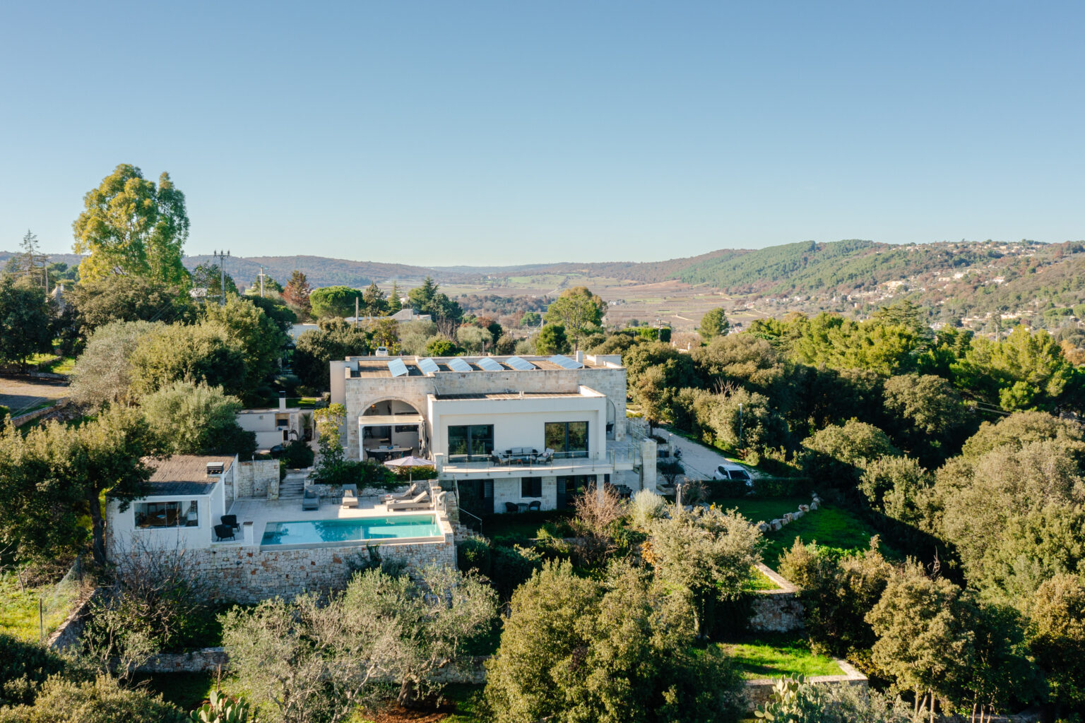 Aerial view of a modern villa with a swimming pool, surrounded by lush greenery and rolling hills in the background, showcasing a tranquil countryside setting. | Only on SopranoVillas