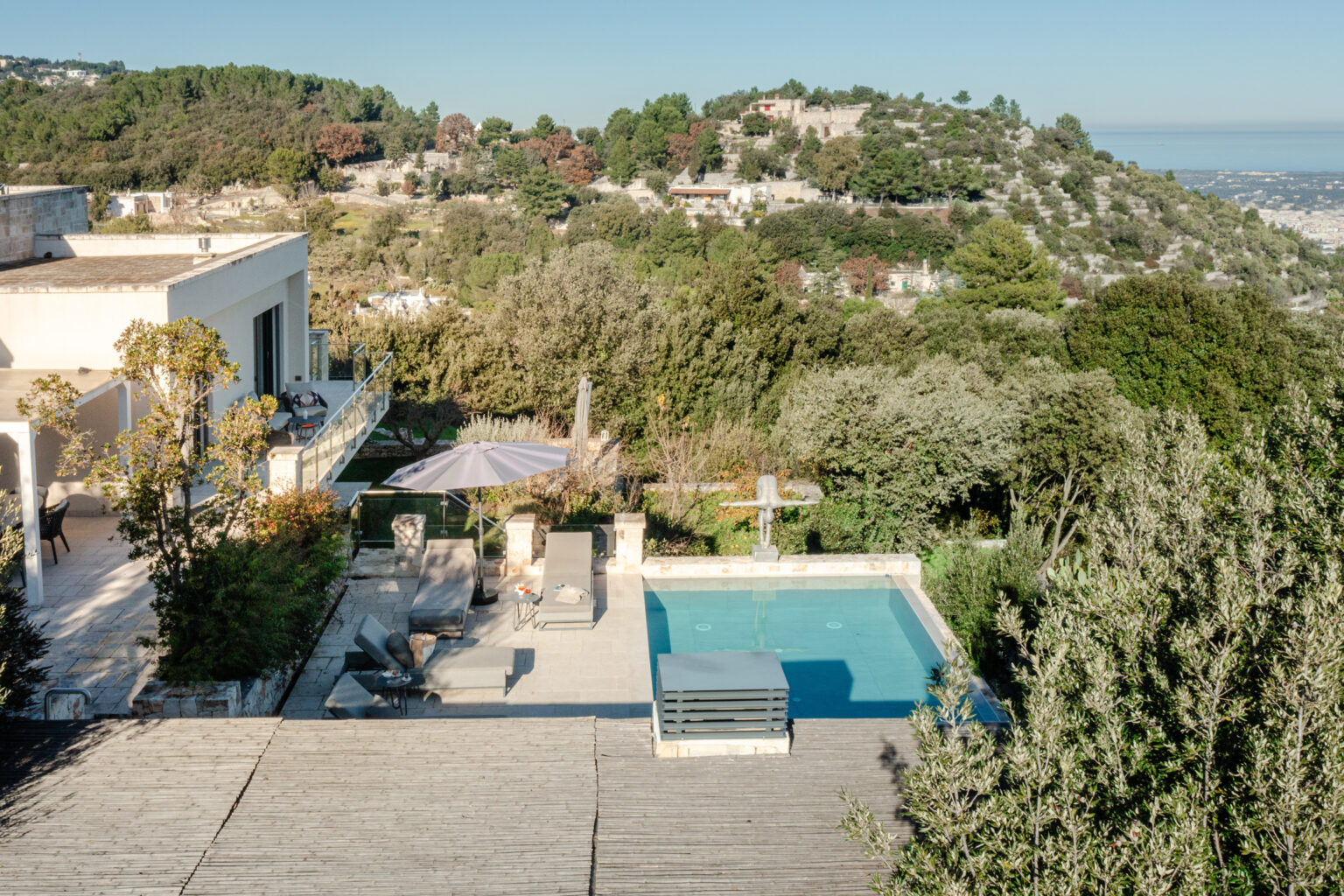 Aerial view of a modern villa with a swimming pool, surrounded by lush greenery and hills. The outdoor area features lounge chairs, an umbrella, and a wooden deck, creating a serene and inviting atmosphere. | Only on SopranoVillas