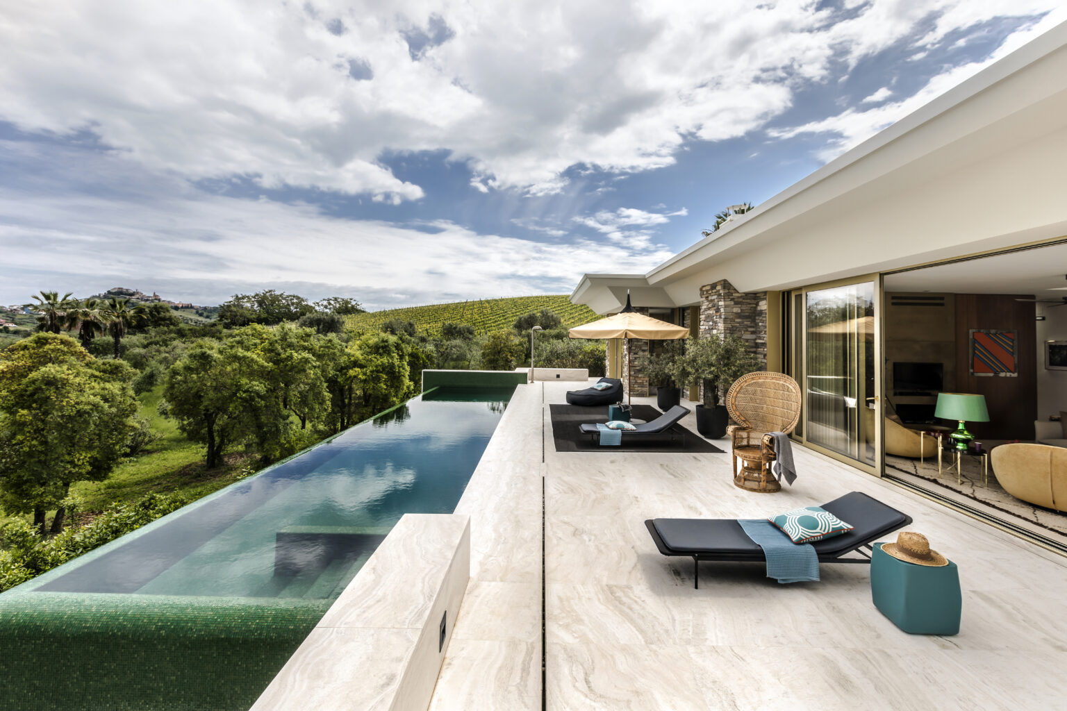 Modern luxury villa with an infinity pool overlooking lush greenery, featuring stylish outdoor furniture and a sun umbrella, set against a dramatic sky. Ideal for relaxation and scenic views. | Only on SopranoVillas
