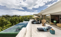 Modern luxury villa with an infinity pool overlooking lush greenery, featuring stylish outdoor furniture and a sun umbrella, set against a dramatic sky. Ideal for relaxation and scenic views.