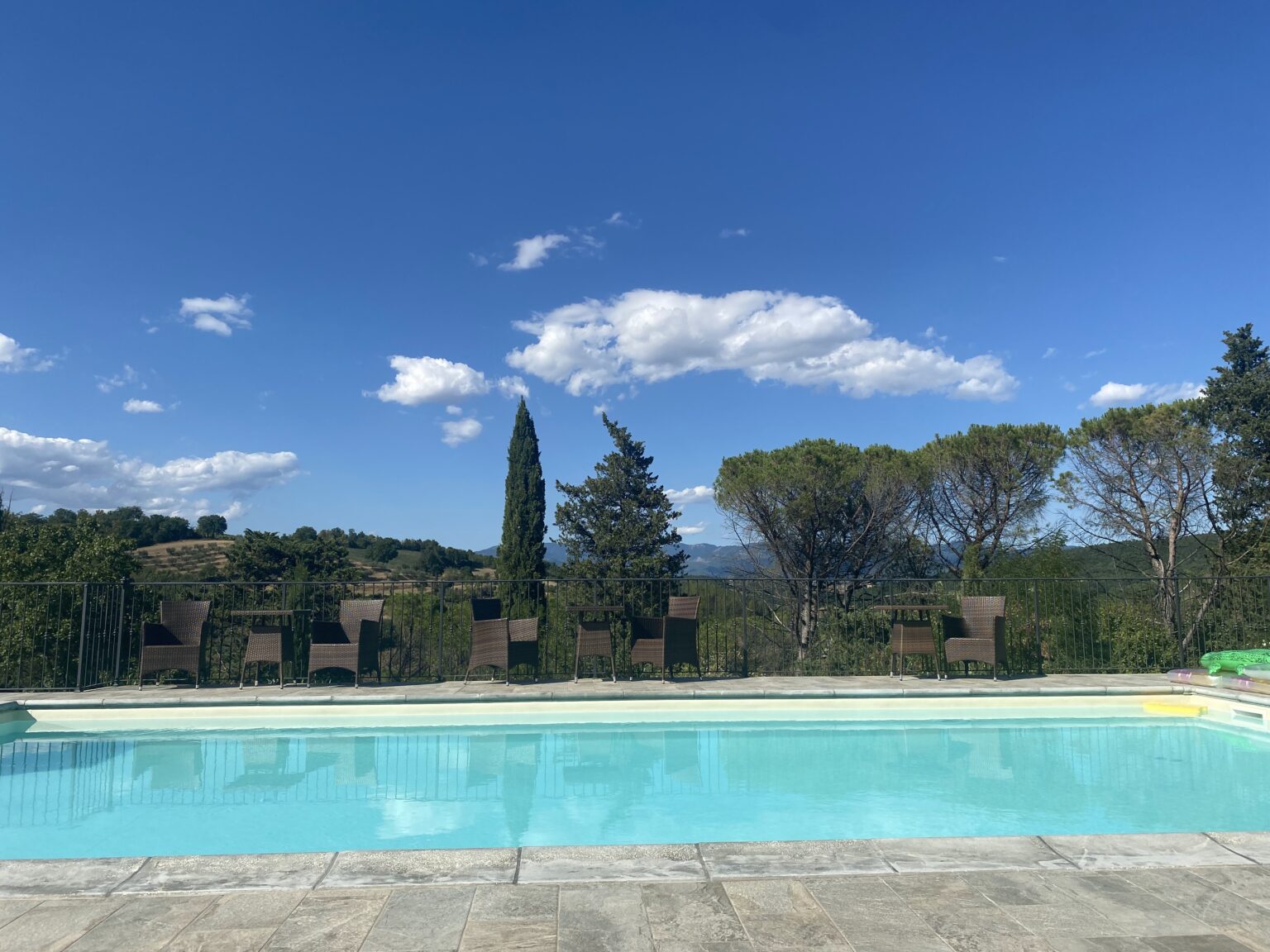 Scenic outdoor swimming pool with clear blue water, surrounded by lounge chairs and lush greenery, under a bright blue sky with fluffy clouds. Ideal for relaxation and leisure in a tranquil setting. | Only on SopranoVillas