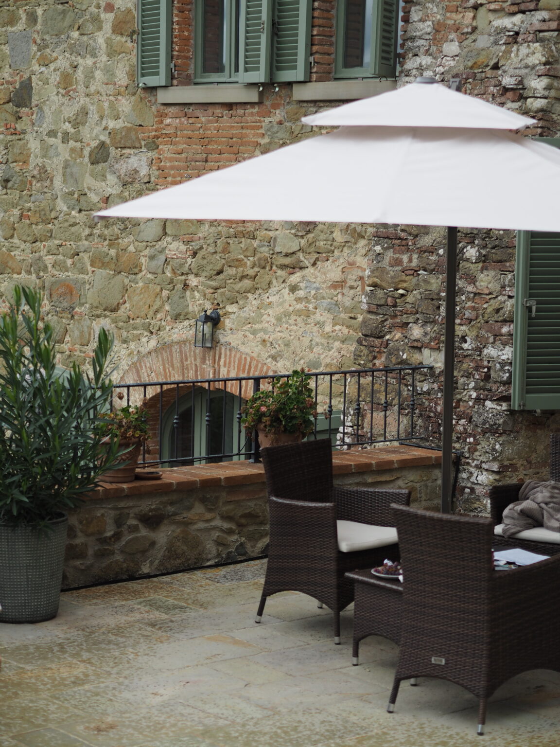 Outdoor patio area featuring a white umbrella over wicker chairs and a table, set against a rustic stone wall with green shutters and potted plants, ideal for relaxation and dining. | Only on SopranoVillas