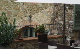 Outdoor patio area featuring a white umbrella over wicker chairs and a table, set against a rustic stone wall with green shutters and potted plants, ideal for relaxation and dining.