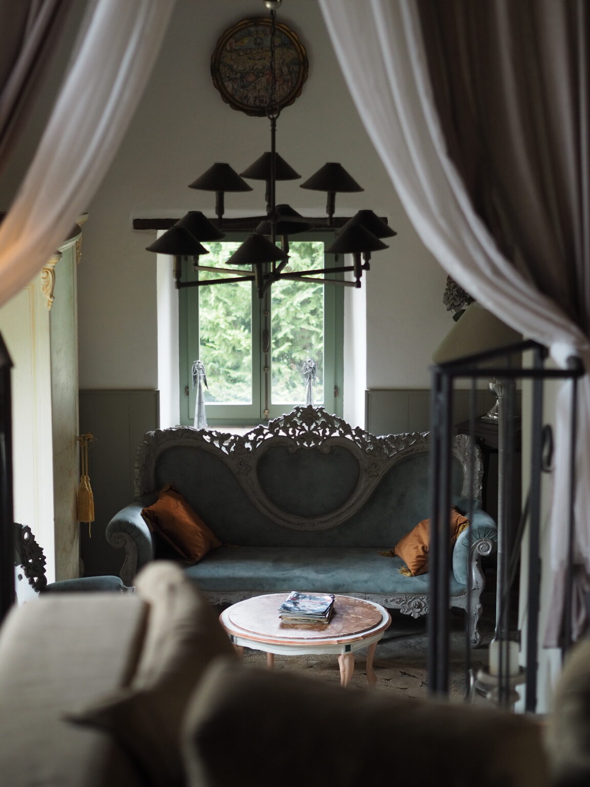 Cozy living room interior featuring a vintage blue sofa with ornate detailing, complemented by orange cushions. A round wooden coffee table holds magazines, while a stylish chandelier hangs above. Natural light filters through a green-framed window, enhancing the inviting atmosphere. | Only on SopranoVillas