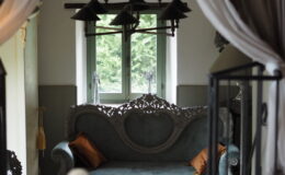 Cozy living room interior featuring a vintage blue sofa with ornate detailing, complemented by orange cushions. A round wooden coffee table holds magazines, while a stylish chandelier hangs above. Natural light filters through a green-framed window, enhancing the inviting atmosphere.