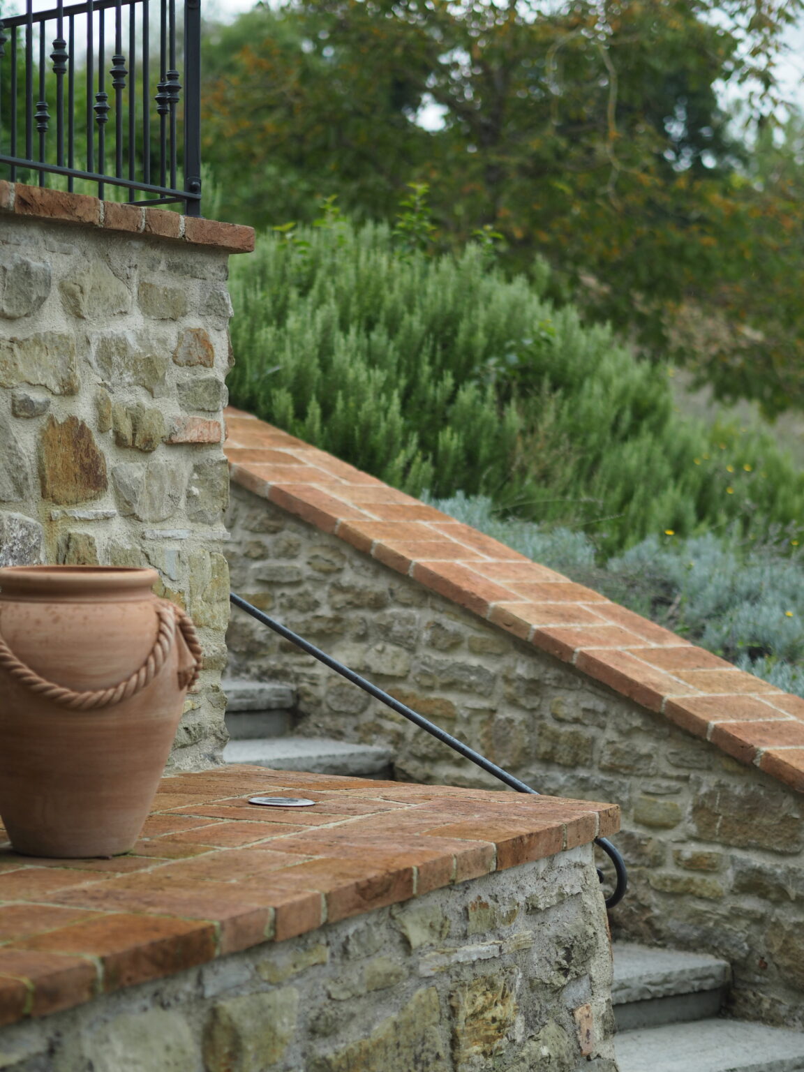 Stone steps lead up to a lush garden, featuring a terracotta pot with a rope handle on a brick surface, framed by greenery and a decorative black railing. | Only on SopranoVillas