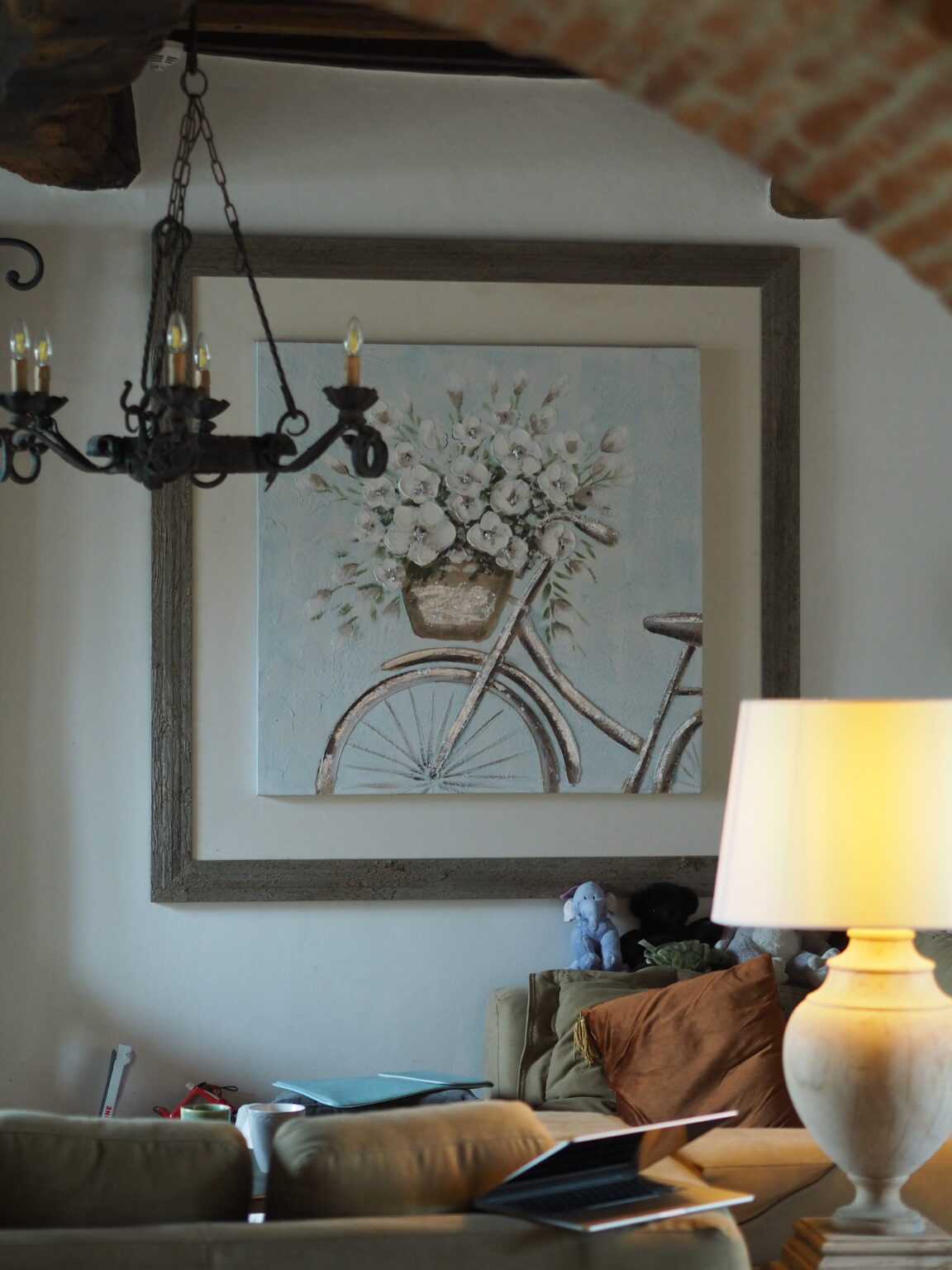 A cozy living room featuring a decorative painting of a vintage bicycle with a flower basket, framed in rustic wood. A chandelier hangs from the ceiling, and a warm lamp illuminates the space, which includes a comfortable sofa with cushions and a laptop on a side table. | Only on SopranoVillas