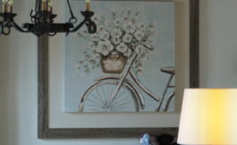 A cozy living room featuring a decorative painting of a vintage bicycle with a flower basket, framed in rustic wood. A chandelier hangs from the ceiling, and a warm lamp illuminates the space, which includes a comfortable sofa with cushions and a laptop on a side table.