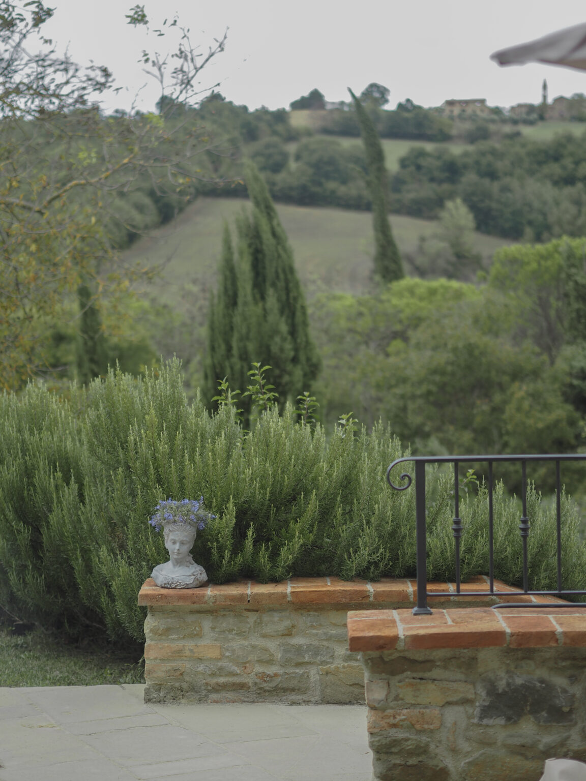 Scenic view of a tranquil garden featuring a stone wall with a bust adorned with flowers, surrounded by lush greenery and cypress trees, set against rolling hills in the background. Ideal for nature lovers and garden enthusiasts. | Only on SopranoVillas