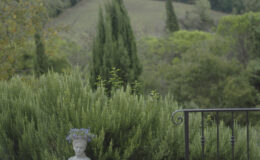 Scenic view of a tranquil garden featuring a stone wall with a bust adorned with flowers, surrounded by lush greenery and cypress trees, set against rolling hills in the background. Ideal for nature lovers and garden enthusiasts.