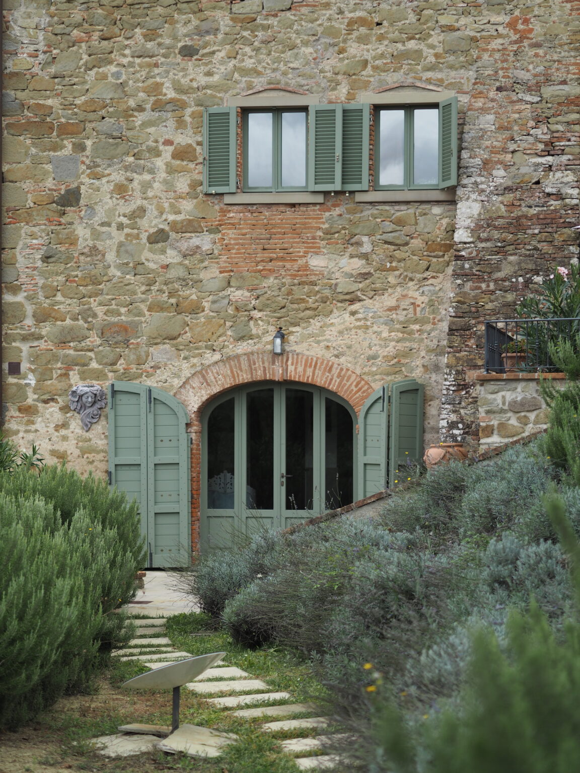 Charming rustic stone house with green shutters and double doors, surrounded by lush lavender plants and a stone pathway. Ideal for showcasing traditional architecture and serene outdoor spaces. | Only on SopranoVillas