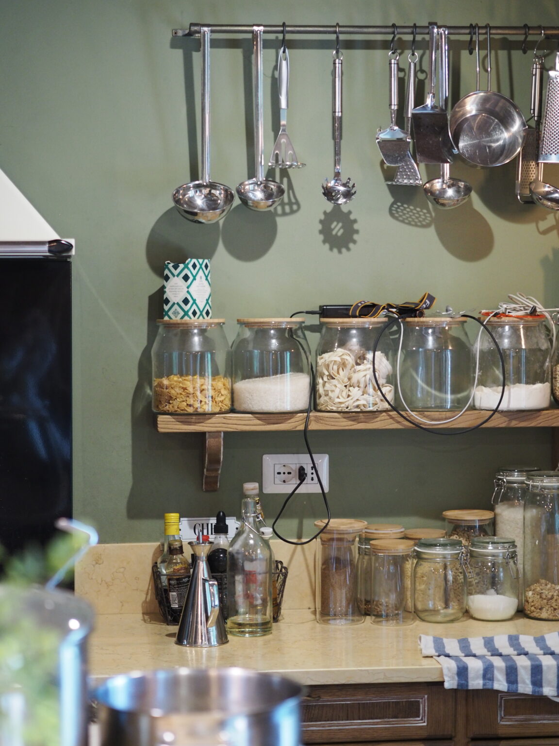 Modern kitchen countertop featuring organized glass jars filled with various ingredients, stainless steel cooking utensils hanging on a rack, and a neutral color scheme with a green wall. | Only on SopranoVillas
