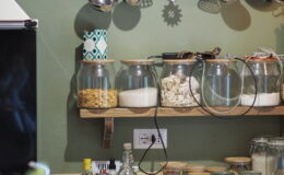 Modern kitchen countertop featuring organized glass jars filled with various ingredients, stainless steel cooking utensils hanging on a rack, and a neutral color scheme with a green wall.