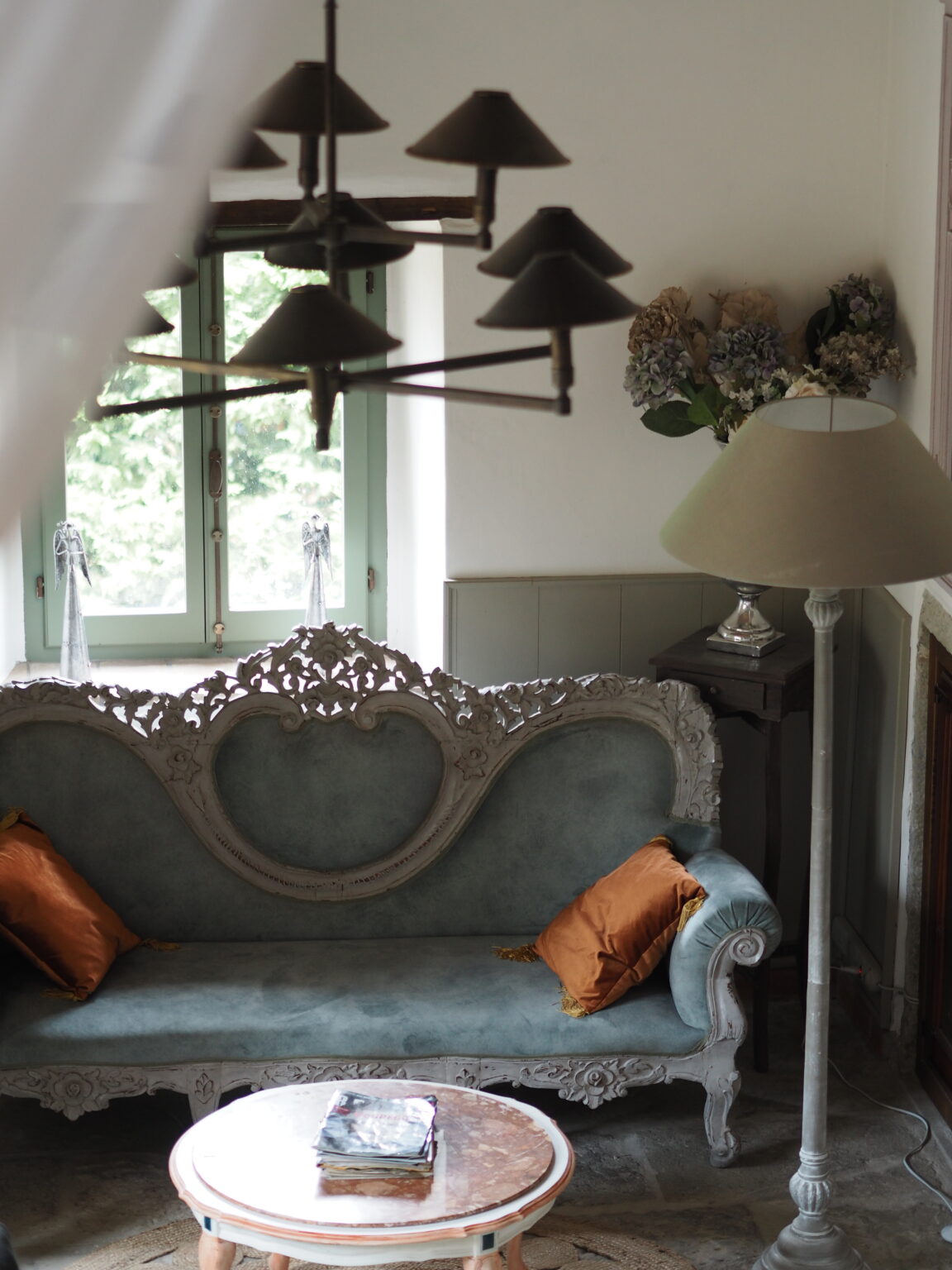Elegant vintage living room featuring a light blue upholstered sofa with ornate detailing and orange decorative pillows, a round coffee table with a marble top, and a stylish floor lamp. Natural light streams in through green-framed windows, enhancing the cozy atmosphere. | Only on SopranoVillas
