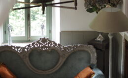 Elegant vintage living room featuring a light blue upholstered sofa with ornate detailing and orange decorative pillows, a round coffee table with a marble top, and a stylish floor lamp. Natural light streams in through green-framed windows, enhancing the cozy atmosphere.