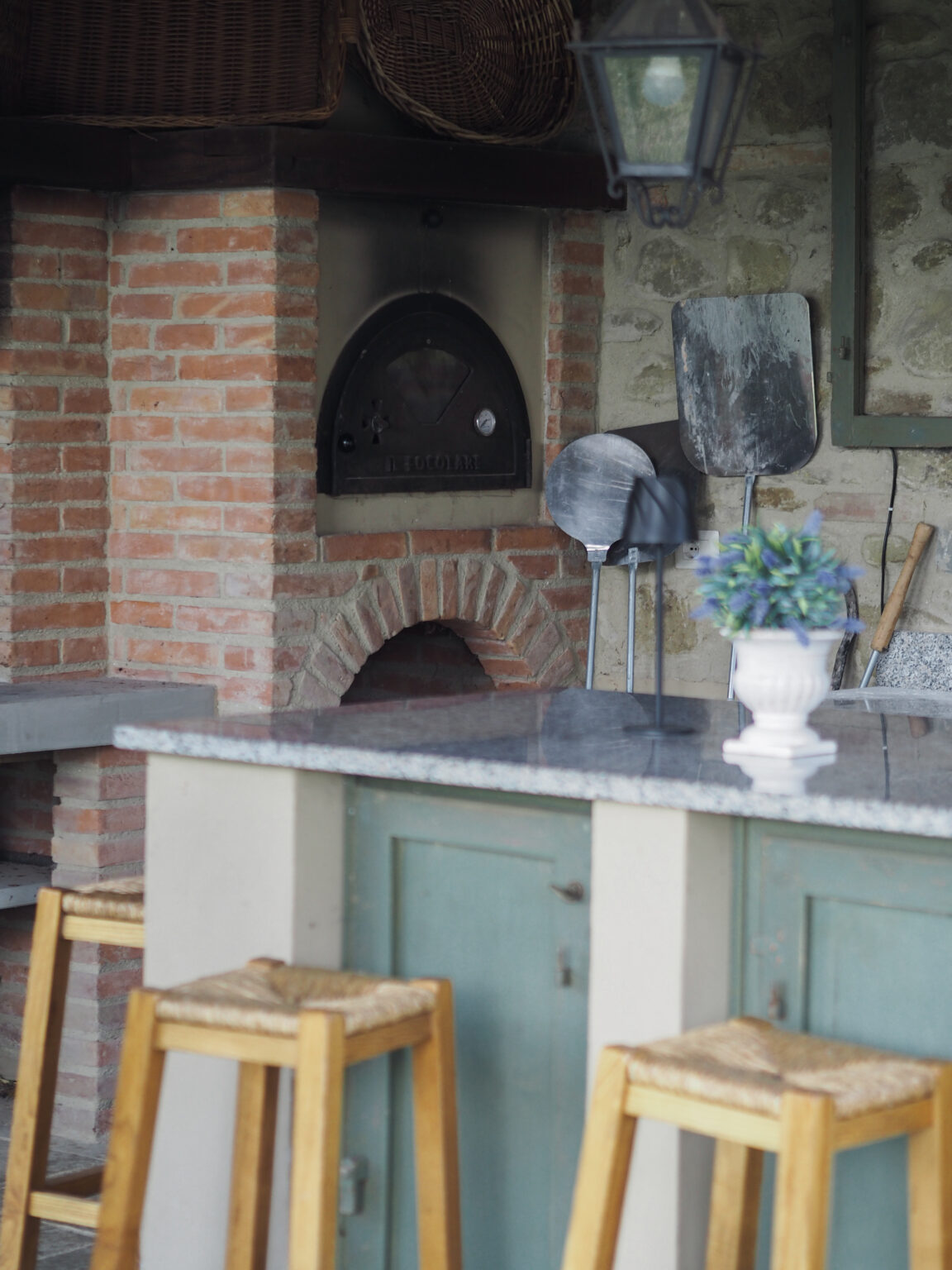 Rustic outdoor kitchen featuring a brick pizza oven, granite countertop, wooden bar stools, and kitchen utensils, surrounded by a charming stone wall. Ideal for outdoor cooking and entertaining. | Only on SopranoVillas