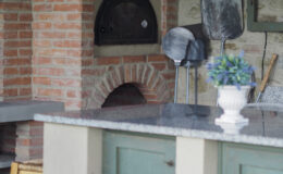 Rustic outdoor kitchen featuring a brick pizza oven, granite countertop, wooden bar stools, and kitchen utensils, surrounded by a charming stone wall. Ideal for outdoor cooking and entertaining.