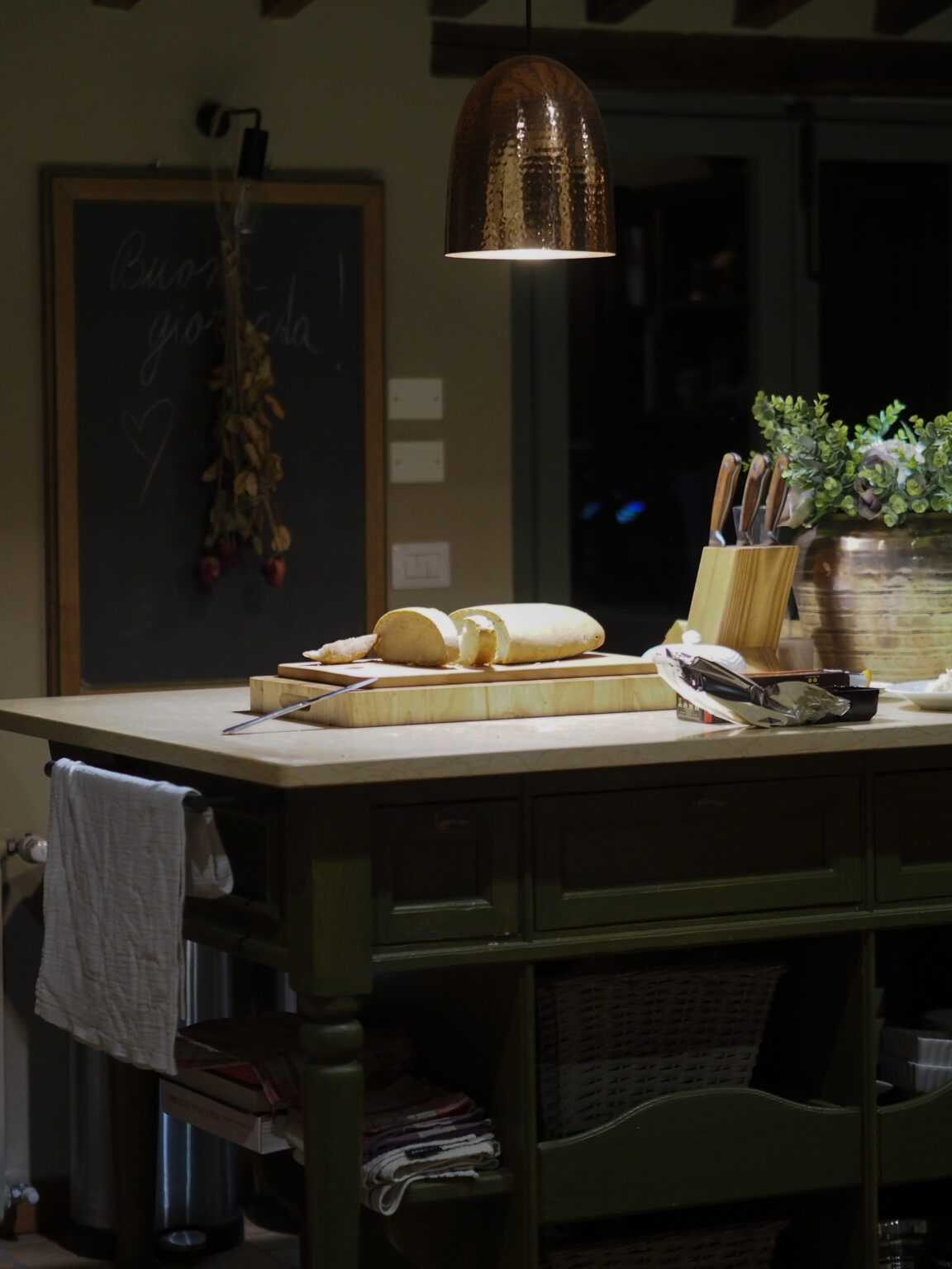 Cozy kitchen interior featuring a wooden island with freshly sliced bread, a copper pendant light, and a chalkboard with handwritten notes. Kitchen utensils and a potted plant add warmth to the inviting atmosphere. | Only on SopranoVillas