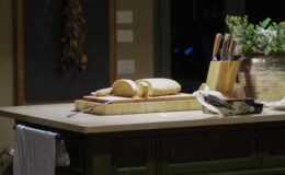 Cozy kitchen interior featuring a wooden island with freshly sliced bread, a copper pendant light, and a chalkboard with handwritten notes. Kitchen utensils and a potted plant add warmth to the inviting atmosphere.