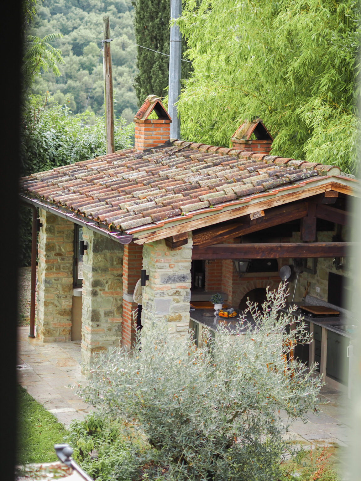 Rustic outdoor kitchen with a tiled roof, stone and brick walls, surrounded by lush greenery and olive plants, set against a backdrop of rolling hills. | Only on SopranoVillas