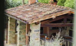 Rustic outdoor kitchen with a tiled roof, stone and brick walls, surrounded by lush greenery and olive plants, set against a backdrop of rolling hills.