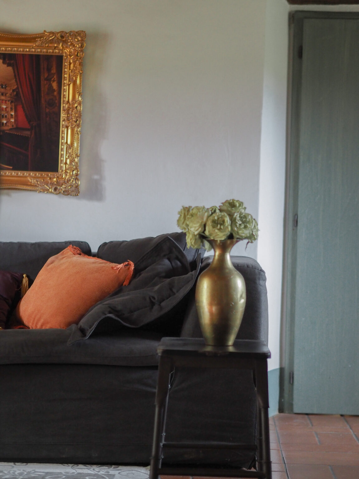 Cozy living room featuring a dark sofa with colorful pillows, a decorative vase with faux flowers on a small black side table, and an ornate gold-framed painting on the wall. A wooden door in the background adds to the inviting atmosphere. | Only on SopranoVillas