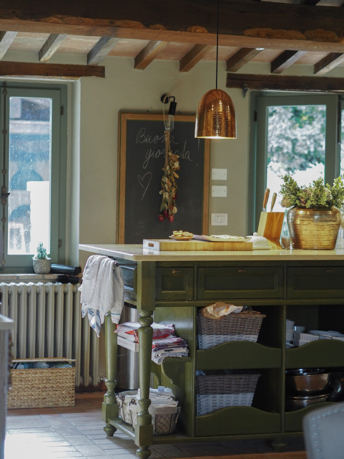 Cozy kitchen interior featuring a green island with storage baskets, a copper pendant light, and a chalkboard with handwritten notes. Natural light streams through windows, highlighting the rustic wooden beams and warm tile flooring. | Only on SopranoVillas