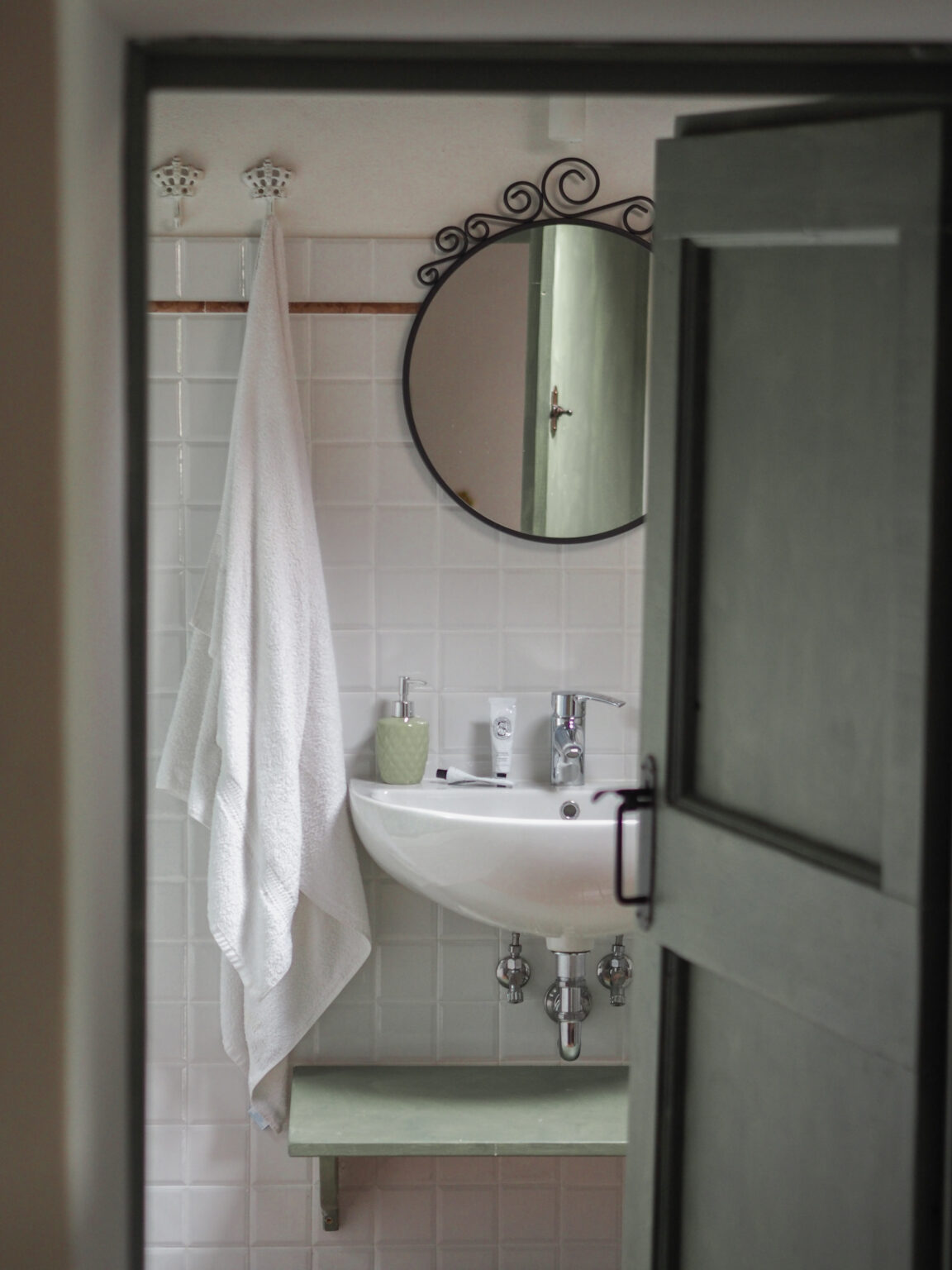 Modern bathroom interior featuring a white sink, a round mirror with decorative frame, and a towel hanging on a hook. A small green shelf holds toiletries, with light-colored tiles creating a clean and inviting atmosphere. The image is taken from an open door, providing a glimpse into the space. | Only on SopranoVillas