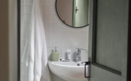 Modern bathroom interior featuring a white sink, a round mirror with decorative frame, and a towel hanging on a hook. A small green shelf holds toiletries, with light-colored tiles creating a clean and inviting atmosphere. The image is taken from an open door, providing a glimpse into the space.