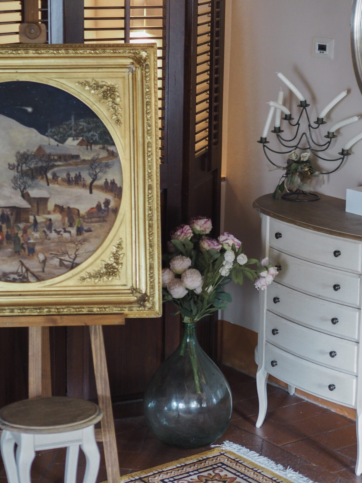 Interior decor featuring a vintage painting in a gold frame on an easel, surrounded by a decorative vase with flowers, a stylish cabinet, and a candelabra, creating an elegant and inviting atmosphere. | Only on SopranoVillas