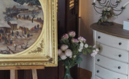 Interior decor featuring a vintage painting in a gold frame on an easel, surrounded by a decorative vase with flowers, a stylish cabinet, and a candelabra, creating an elegant and inviting atmosphere.