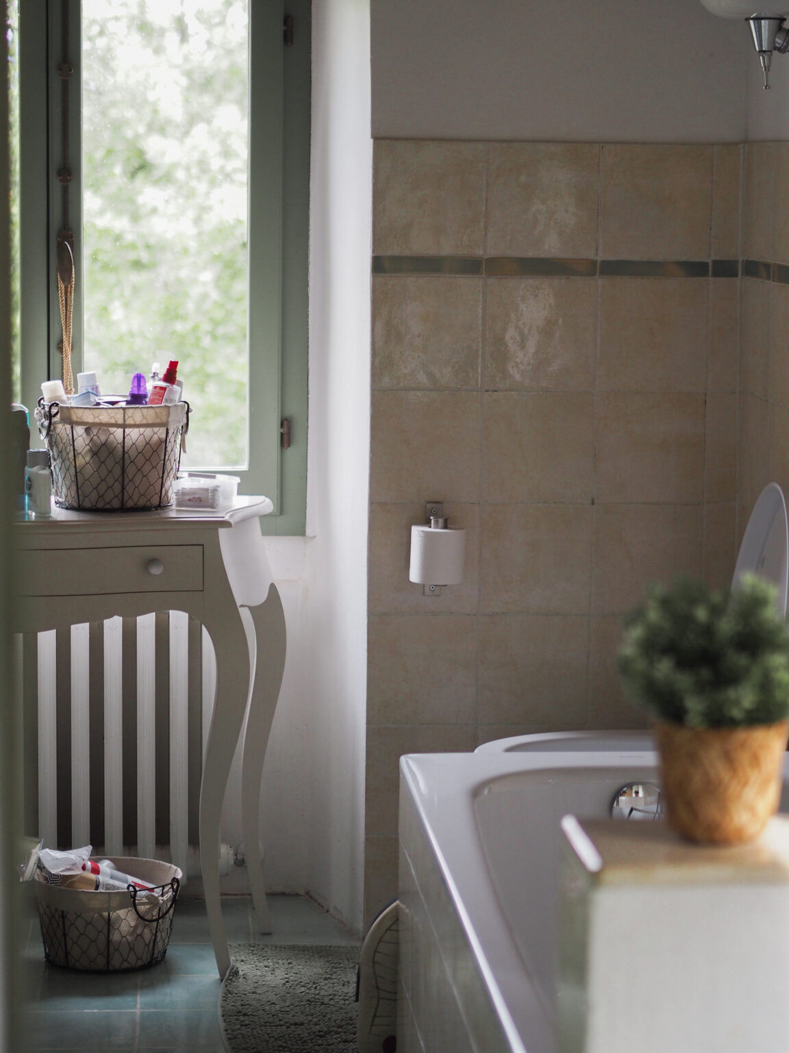 Bright and airy bathroom featuring a bathtub, a stylish white vanity with storage baskets, and a cozy green rug. Natural light streams through a large window, illuminating the space adorned with toiletries and a small potted plant. | Only on SopranoVillas