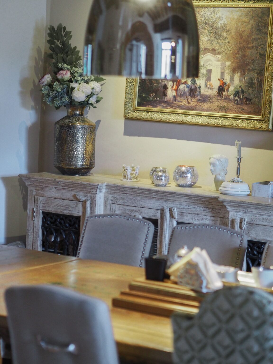 Elegant dining room featuring a rustic wooden table, plush chairs, and a decorative sideboard adorned with a vintage vase of flowers, crystal candle holders, and an ornate painting in a gold frame, creating a warm and inviting atmosphere. | Only on SopranoVillas