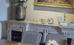 Elegant dining room featuring a rustic wooden table, plush chairs, and a decorative sideboard adorned with a vintage vase of flowers, crystal candle holders, and an ornate painting in a gold frame, creating a warm and inviting atmosphere.